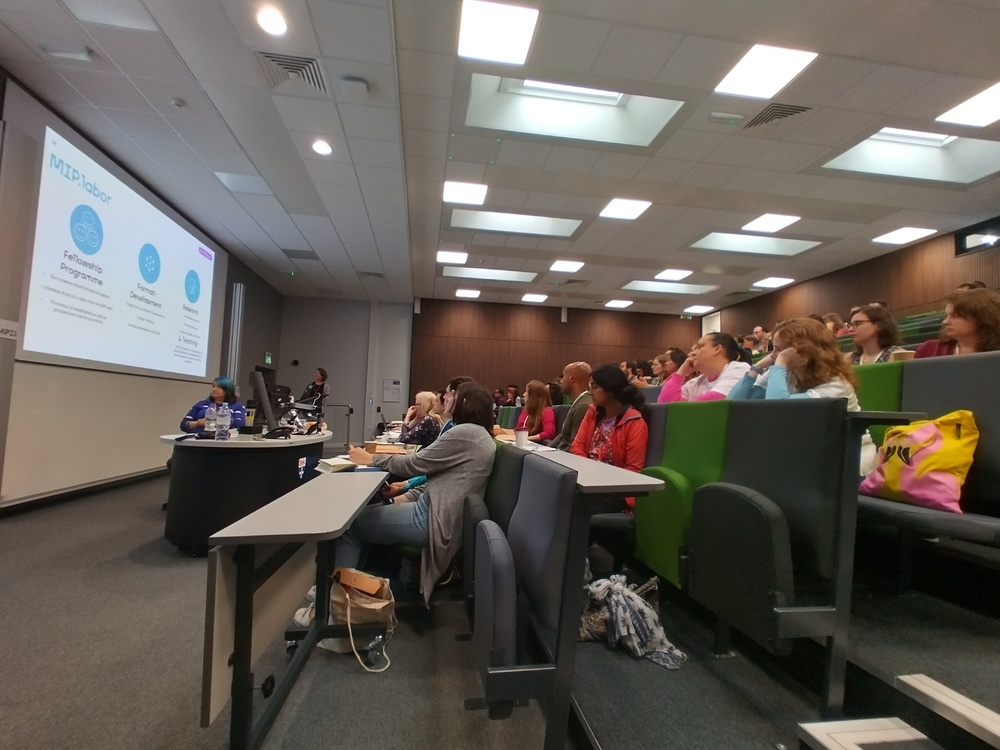 A lecture theatre with banked seating looking at a presenter stood in front of a large projector screen.