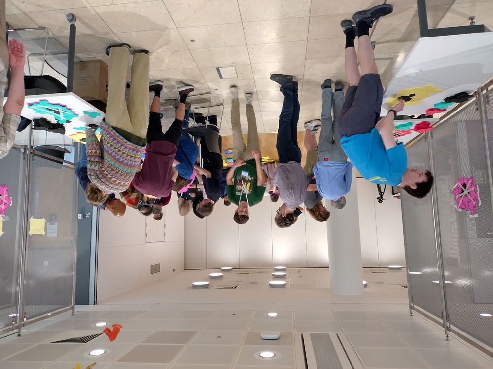 People stood in an open public area around tables and chatting with each other. There are some people seated on chairs.